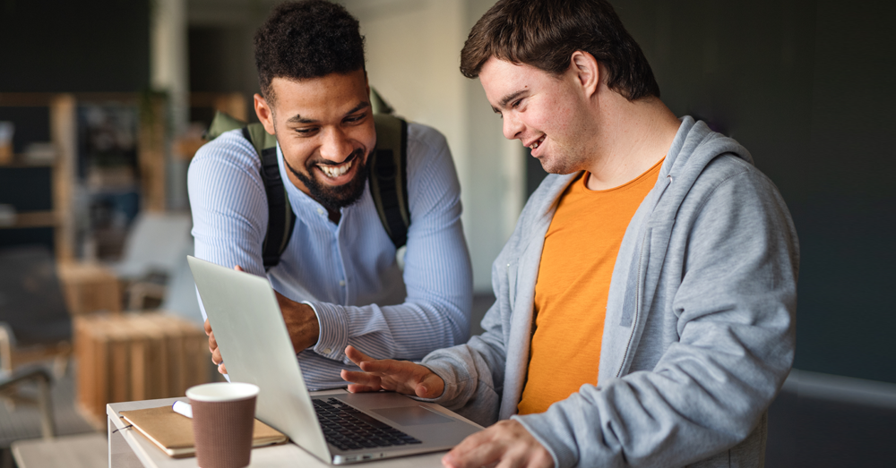 Auf dem Bild siehst du zwei junge Männer.
Sie gucken gemeinsam auf einen Laptop.
Die beiden Männer lachen.